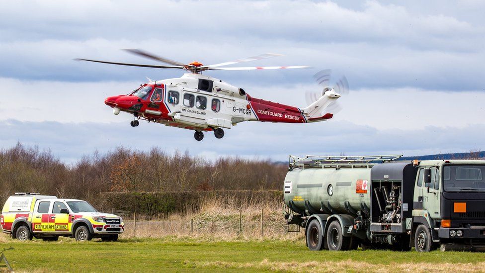 A red and white coastguard helicopter comes in to land next to a fuel truck and airport pick up truck