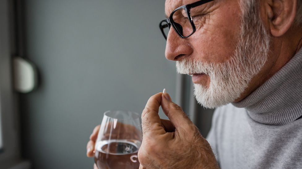 A man with a white beard holds a glass of water while taking a pill
