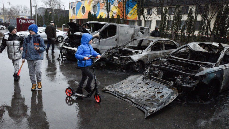Kids walk past next to destroyed cars after a missile attack