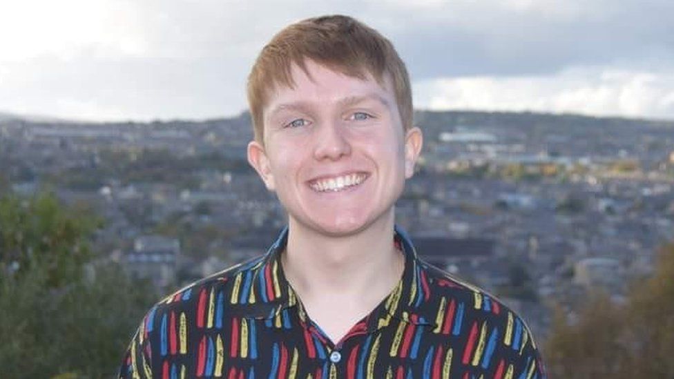 Joe Thompson. Joe is a white man with short strawberry-blonde hair and blue eyes. He wears a black shirt with yellow, red and blue stripes and smiles at the camera. He's pictured outside on a hill with trees and a town or city behind him.