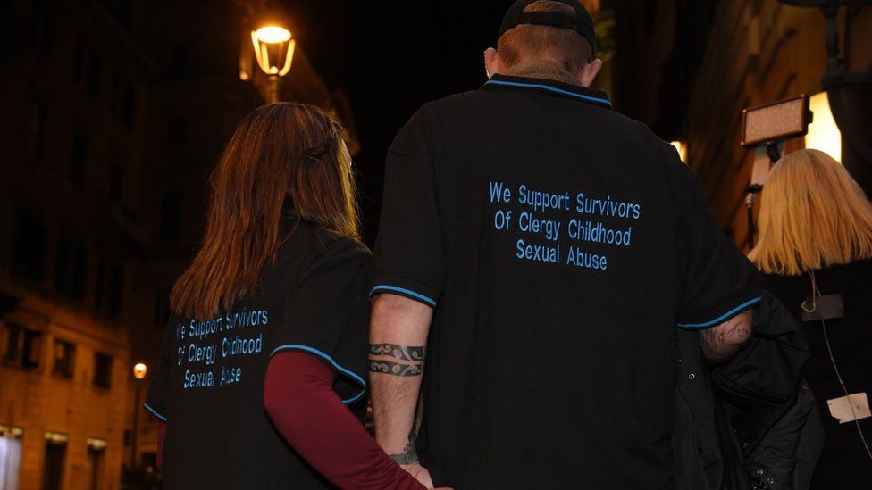 A survivor of child abuse, and his wife, hold hands while wearing T-shirts that read: "We support survivors of clergy childhood sexual abuse"
