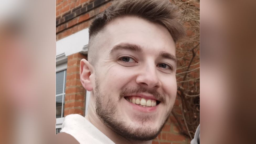 A man smiles at the camera - he's got brown eyes and shortish brown hair on top of his head. The sides of his head are closely shaved and he has a short beard and moustache. He's stood in front of a redbrick house. We can just see the brick work and the white frames around the windows.