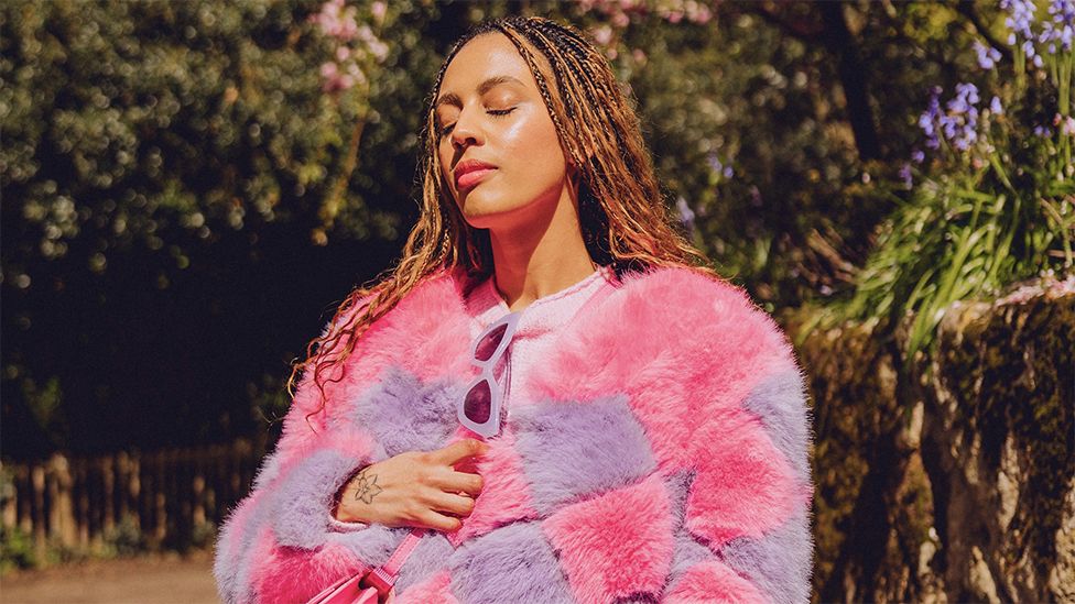 Demi has her eyes closed, as the sun shines on it. She is wearing a pink and purple fur coat, with white sunglasses tucked into the top. Her right hand is towards the middle of her coat. The background has lots of green trees and bushy branches.