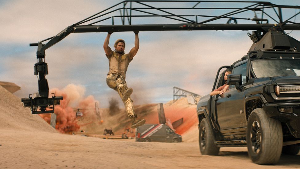 Ryan Gosling is Colt Seavers in The Fall Guy. Gosling is a white male actor in his early 30s. He has fair hair and a muscular physique. In the shot from the film, he hangs from a camera rig that's attached to a black 4x4. The vehicle is in motion, filming an explosion scene in a desert where in the background a helicopter can be seen being engulfed in the smoke.