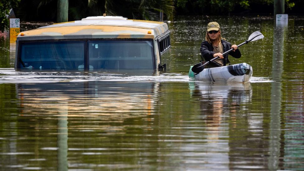 Flooded street with canoeist paddling past a bus with water up to its windscreen
