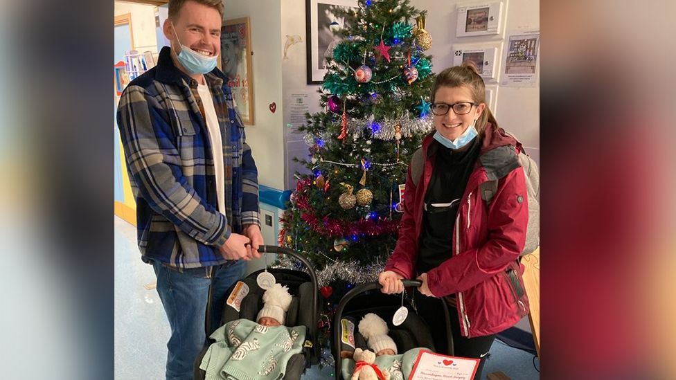 Ruth and Gavin Coleman standing in front of a Christmas tree smiling, each holding one twin in a baby carrier