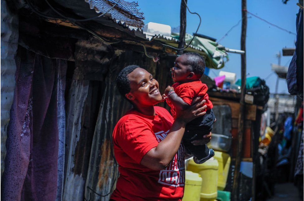 Woman carrying her child outside in Mathare slum. She is smiling, against a backdrop of blue skies and homes.