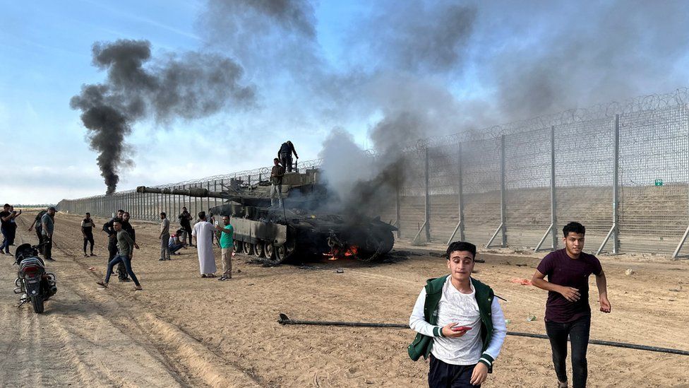 Palestinians react as an Israeli tank burns on the Israeli side of Israel-Gaza perimeter fence (7 October 2023)