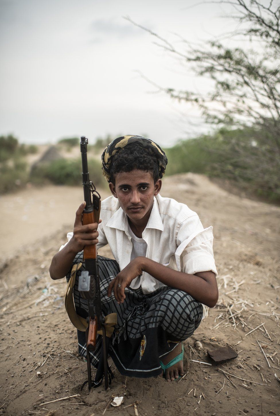 A young man in a Yemen conflict zone crouches whilst holding a gun