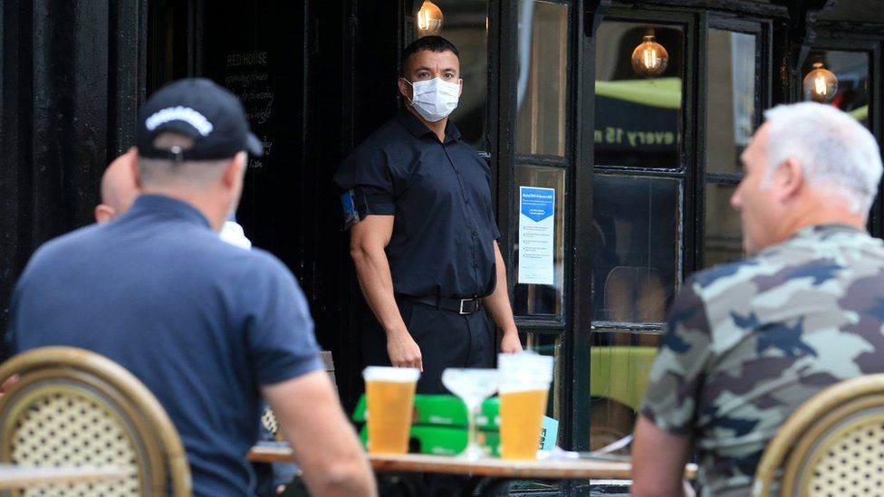 A doorman wearing PPE (personal protective equipment), of a face mask or covering as a precautionary measure against spreading COVID-19, stands on duty as customers sit with their drinks at a re-opened pub in Newcastle