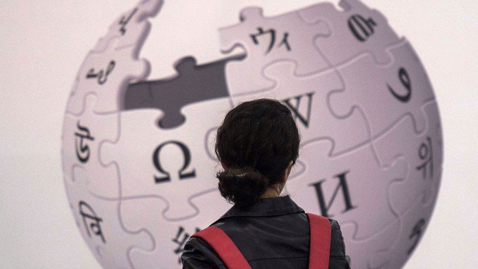 A woman stands with her back to the camera, in front of an enormous Wikipedia logo of various jigsaw pieces with different symbols forming a globe