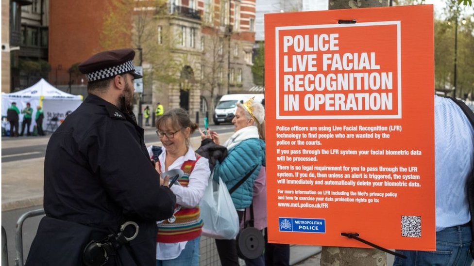 Полицейский стоит рядом с табличкой с надписью: "Полиция распознает лица в режиме реального времени в операция'
