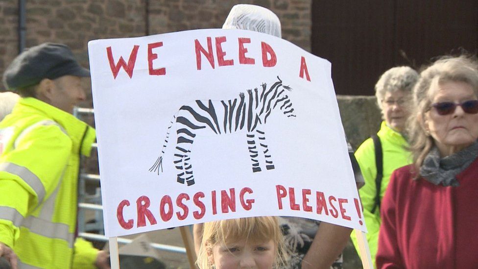 Girl carrying a sign saying 'we need a crossing please'