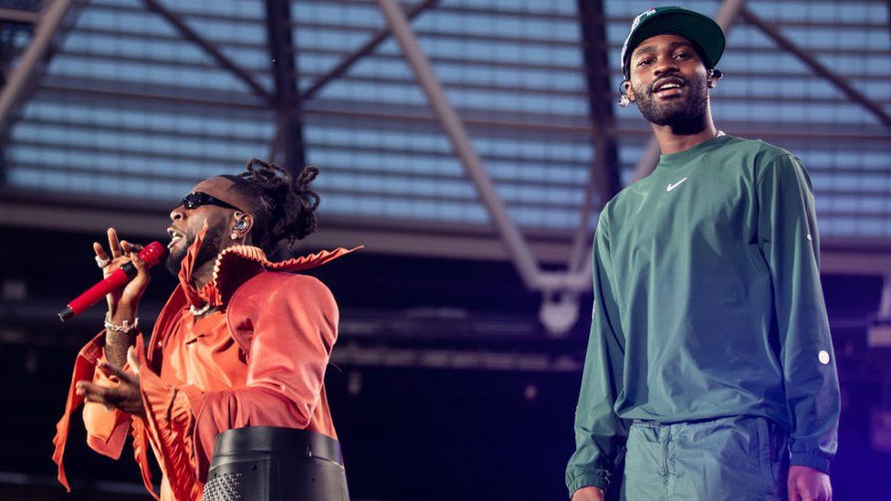 Burna Boy and Dave on stage at the London Stadium. Burna Boy is wearing an orange jumpsuit and holding a red microphone and looking at the crowd as he sings. Dave is on his right and is wearing a green tracksuit Nike set and a cap and looking at the camera.