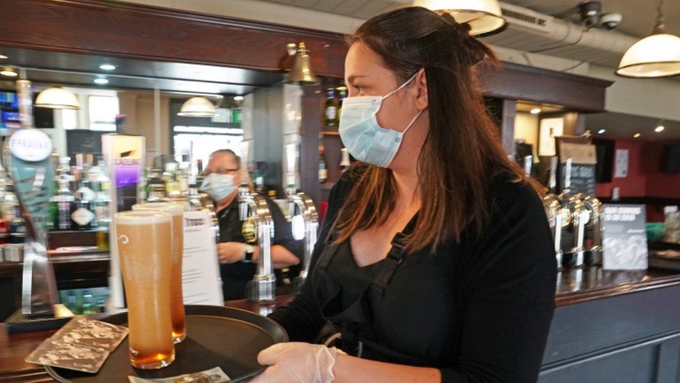 A waitress wearing a face mask carrying a tray holding two pints of beer