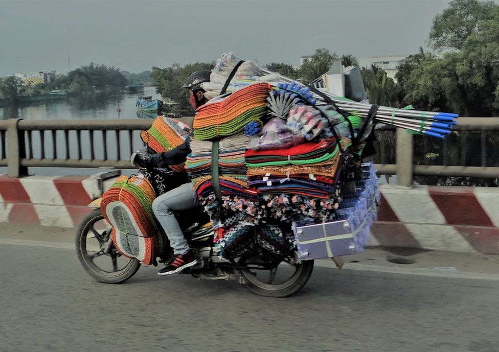 A moped is packed with bath mats, boxes and mops, a driver is crammed in as he drives on