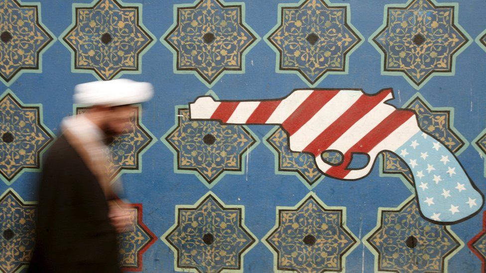 An Iranian cleric walks past a mural on the wall of the former US embassy in Tehran (11 February 2007)