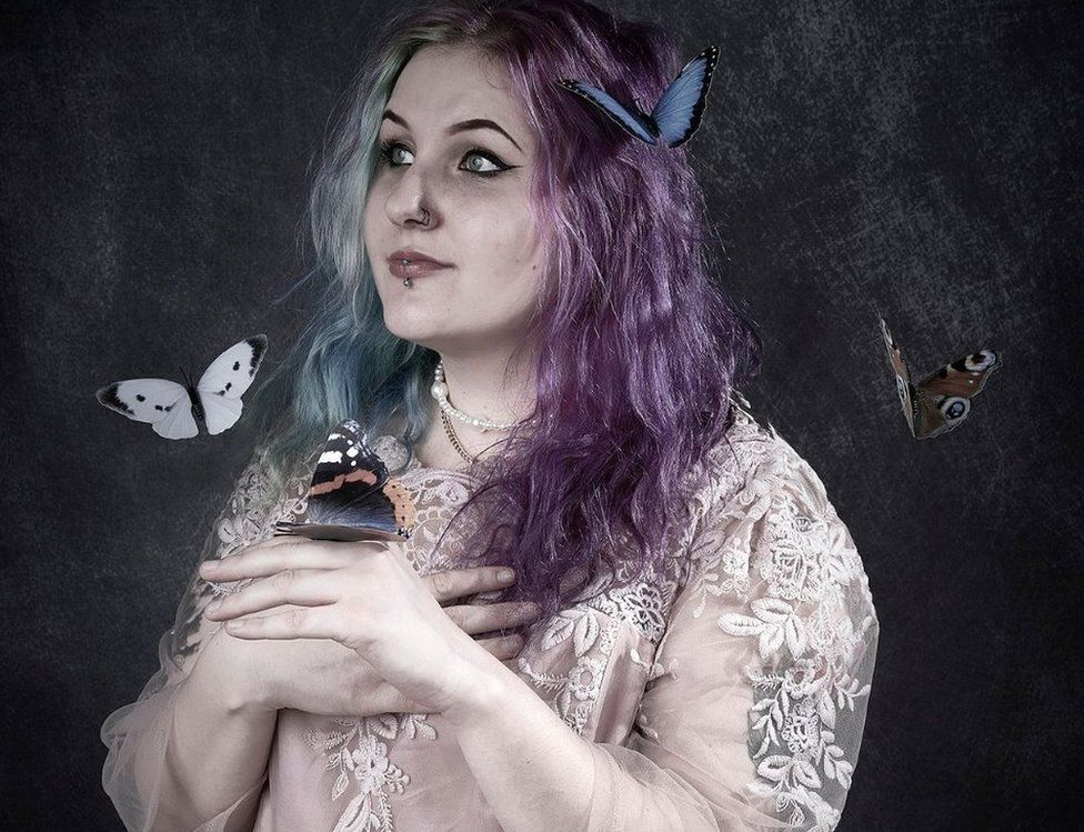 A young woman with purple and green hair and wearing a white dress plays with butterflies superimposed around her