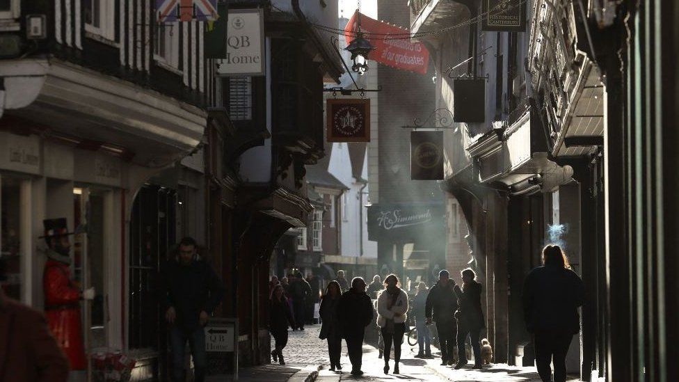 A narrow street in Canterbury, with people walking down it and shop signs hanging overhead