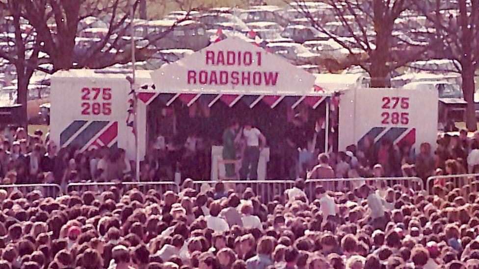 Radio 1 branded stage built onto the side of a lorry, with huge crowd in front and car park behind