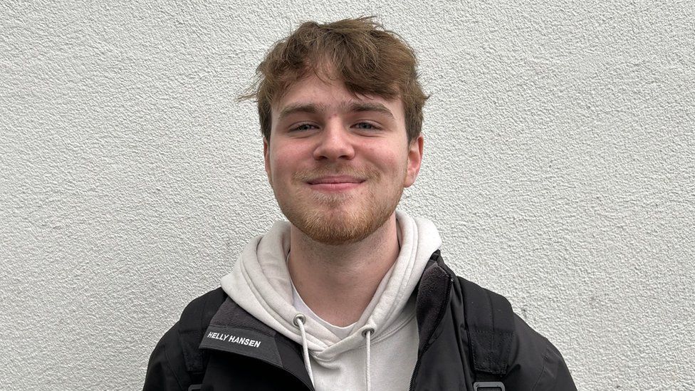 Luke Saklatvala. Luke is a 17-year-old white man with short brown hair and a brown stubbly beard. He has blue eyes and looks happily at the camera, wearing a black jacket over a white hoody. He's pictured outside in front of a white wall.