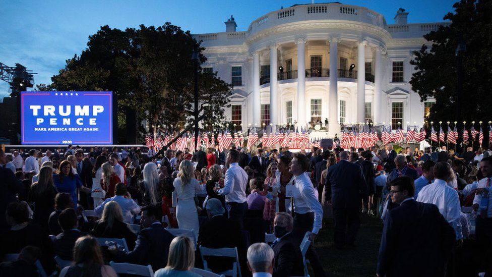 The White House is illuminated by lights. In front is a row of American flags, a Trump/Pence campaign poster and a crowd.