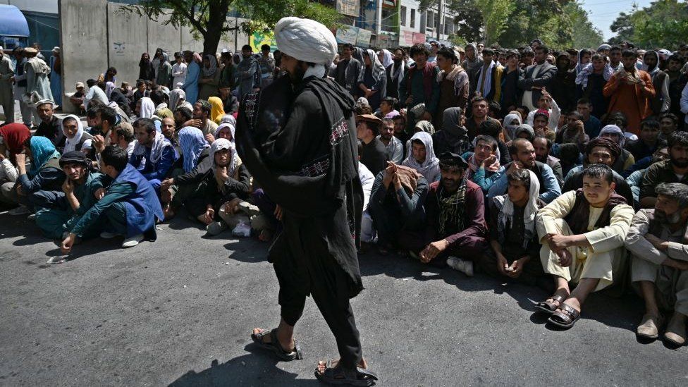 A Taliban fighter walks in front of people sitting along a road outside a bank waiting to withdraw money at Shar-e-Naw neighborhood in Kabul.