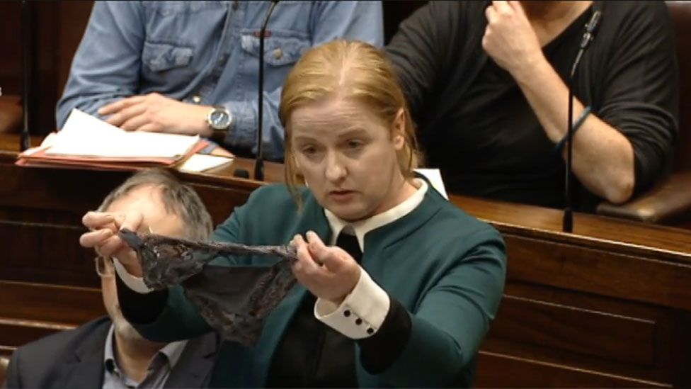 Ruth Coppinger, wearing a blazer, holds out a dark pair of underwear while standing in the dark wooden rows of seating the Irish parliament, Dáil Éireann.