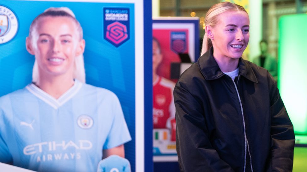Chloe Kelly standing in front of a board showing a life-sized Panini sticker of herself. The sticker shows her against a blue background with the Manchester City badge in one corner and the Barclays Super League logo in the other. Chloe's wearing the sky blue home kit of Manchester City, which has the Etihad Airways logo across the chest. The real-life Chloe is wearing a black bomber jacket, her hands clasped at waist level as she talks to someone off-camera.
