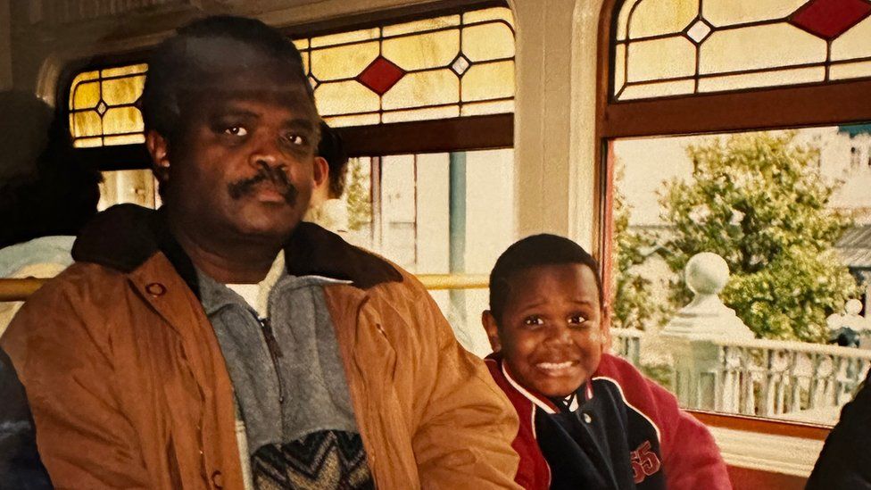 A slightly faded photograph of a father and his young son sitting next to each other on a bus. Both are of African descent. The father, who wears a tan jacket over a grey zip-up top, has a neutral expression. Meanwhile his son, who wears a blue and red USA college-style sports jacket, flashes a cheeky, beaming smile at the camera. In the background ornate fences and pruned trees suggest they are in a tourist spot on a family day out.