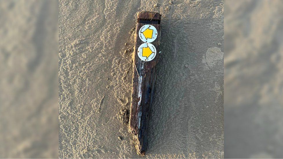 A waterlogged footpath signpost from Shropshire which has been broken off at the bottom and is lying in the sand