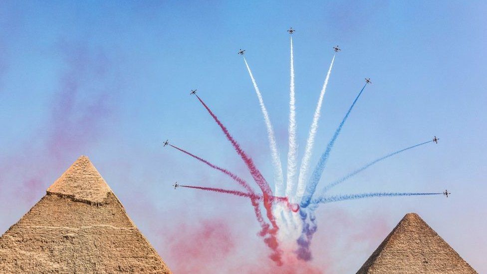 Aircraft are flying in different directions next to pyramids. There is a blue sky, and the aircraft are ejecting red, blue and white colours.