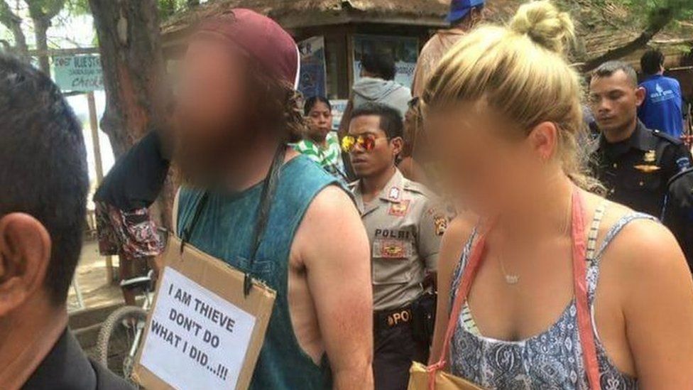 Western man and woman being paraded by Indonesian police and security guards with a sign around their necks reading "I am thieve (sic) don't so what I did...!!!"