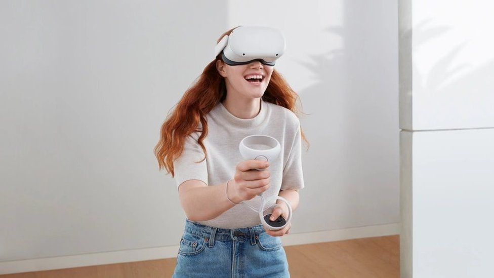 A red-haired woman wears the Oculus Quest 2 headset in white, holding two controllers
