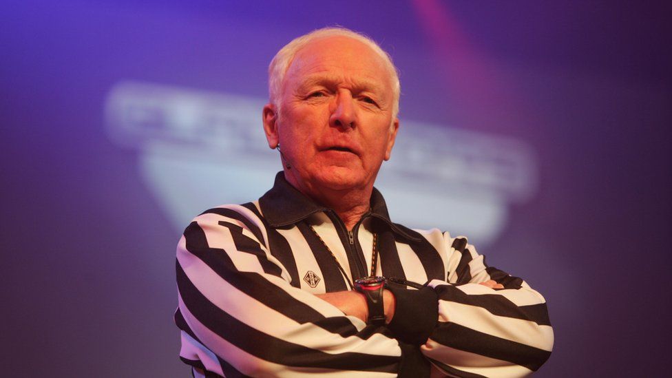 Man with short white hair wearing black and white striped top standing in front of Gladiators logo
