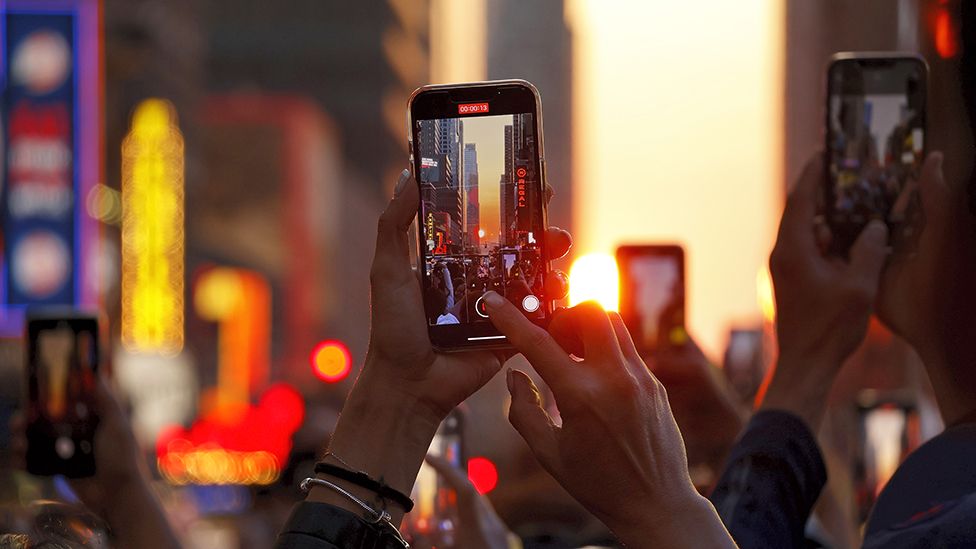 People stand on 42nd Street in Times Square as they photograph the second evening of the Manhattanhenge sunset on May 30, 2023, in New York City