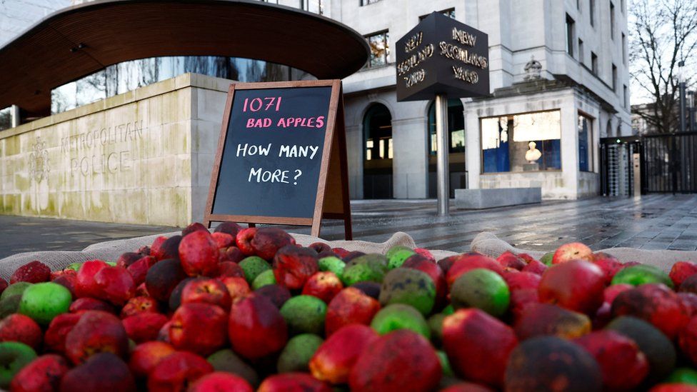 Image showing "rotten" green and red plastic apples in a crate, and a sign saying "1,071 bad apples, how many more?" outside New Scotland yard.