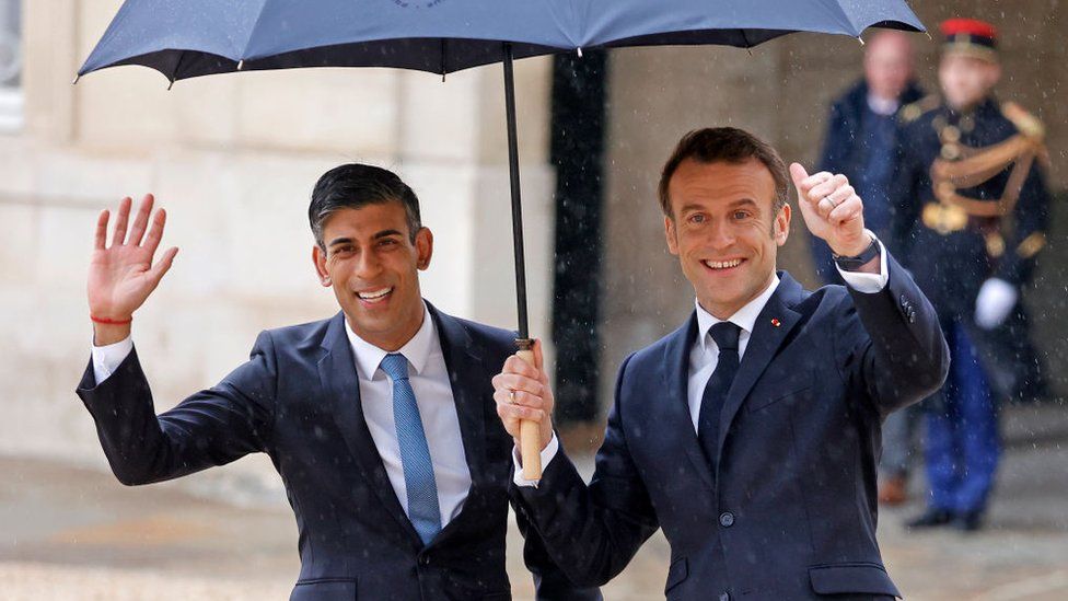 Rishi Sunak and Emmanuel Macron smiling and gesturing to onlookers under an umbrella