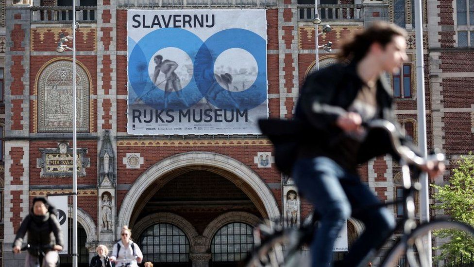 Люди катаются на велосипедах 12 мая 2021 года в Амстердаме возле Рейксмузеума с плакатом выставки «Рабство». -