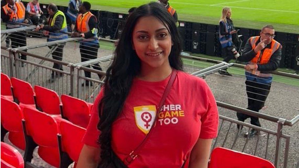 Roopa Vyas stood smiling for the camera at a football stadium. Behind her there are red plastic seats and a few stewards stood in orange hi-vis jackets as well as a few photographers and at the top of the photo you can just make out the green grass from the start of the football pitch. She is an Asian woman with long dark hair and is wearing a red T-shirt with "Her Game Too" written on it as well as their logo.