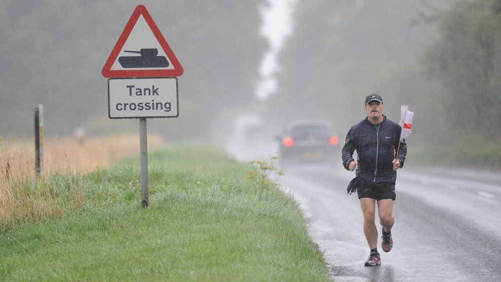 Eddie Izzard running in rain, holding England flag, past sign reading "Tank Crossing"