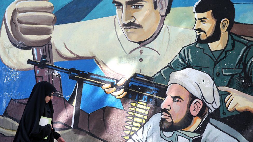 Woman wearing veil and sunglasses walks past mural depicting three men with guns