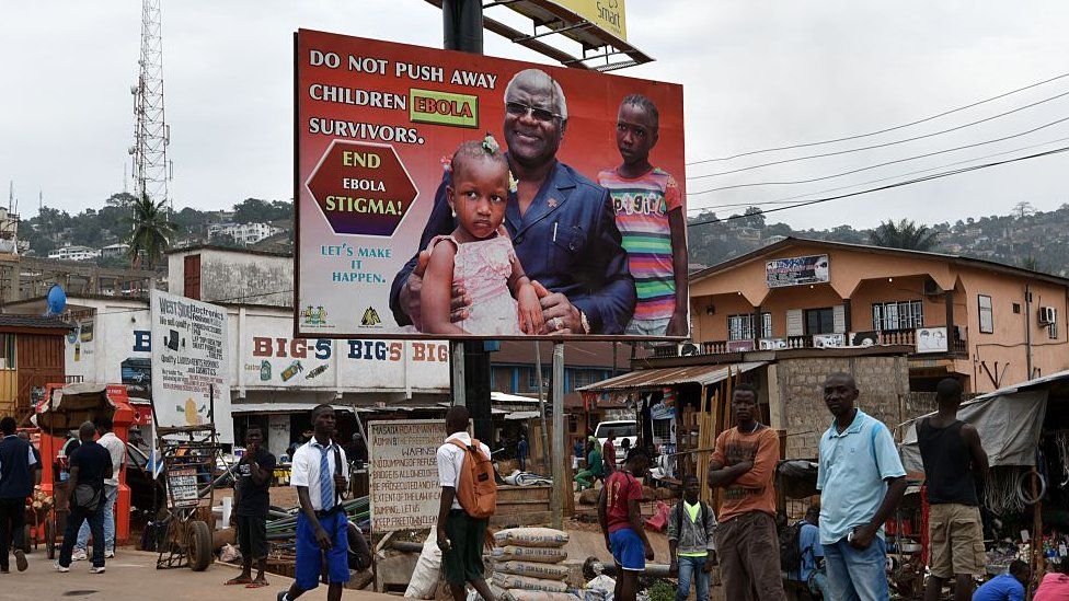 People walk under an Ebola information board featuring Sierra Leone's president Ernest Bai Koroma