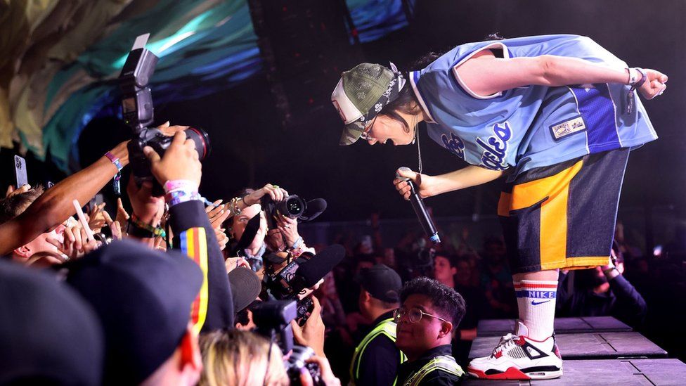 Billie Eilish performing at Coachella Festival. Billie is a 22-year-old white woman with shoulder-length dark hair. She is pictured on stage, bending towards the crowd. She wears blue and yellow sports shorts, matched with a blue sports jersey, white sports socks and white sneakers with red trim. She wears a green and white cap over a blue bandana and holds the microphone in her right hand while her left is behind her back. A line of security officers separates her from the crowd, which is otherwise very close. People in the crowd hold phones and cameras up to capture her performance.