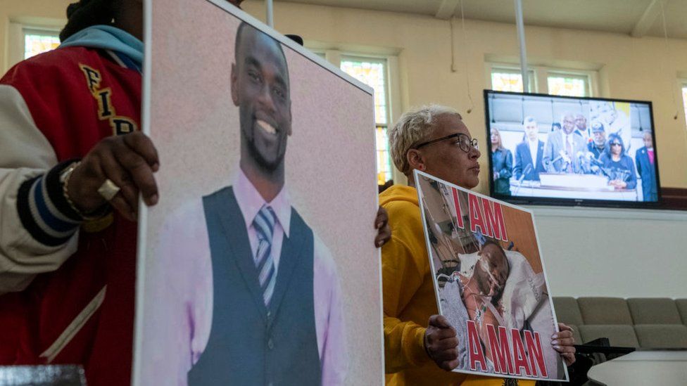 Активисты держат плакаты с изображением Тайра Николса в роли адвоката Бена Крампа, выступающего на мониторе во время пресс-конференции