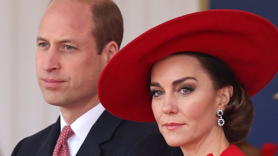 Prince William and Kate Middleton looking into the distance. William is wearing a suit, while the princess is wearing a red had and earings