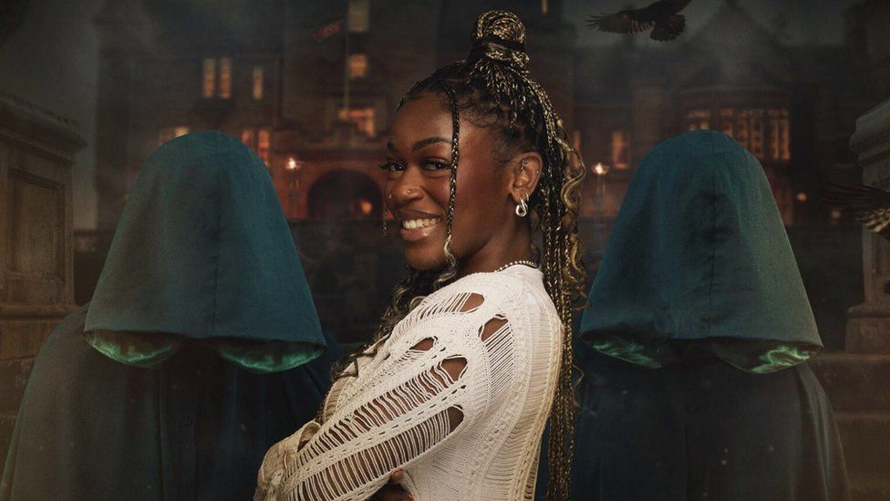 Jasmine Boatswain in a promotional photo for The Traitors. Jasmine is a 26-year-old black woman with long braids. She wears a long-sleeved crochet cream top. She stands facing right, arms folded, and turns her head to smile at the camera. She is flanked by two cloaked traitors in front of the Scottish castle where the series is filmed and flames have been added for dramatic effect