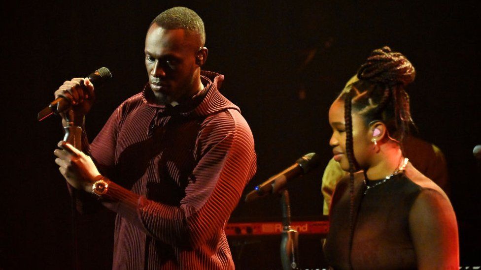 Stormzy and Debbie perform together onstage. Stormzy is wearing a red hoodie, in a ribbed texture. He holds the microphone with both hands. Debbie wears her hair up, she's wearing a sparkling necklace and smiling, looking out to the audience.