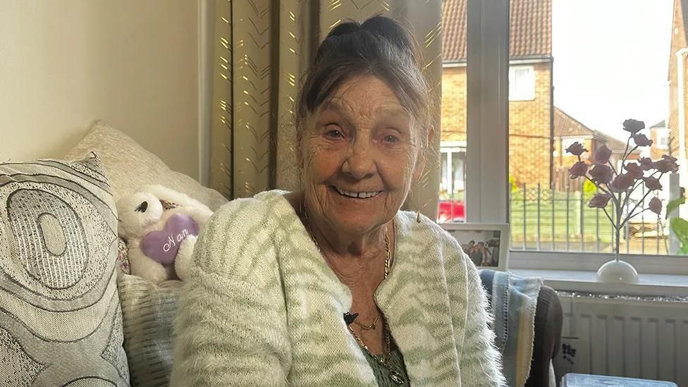 An elderly woman sits on her sofa at home. She is smiling at the camera.