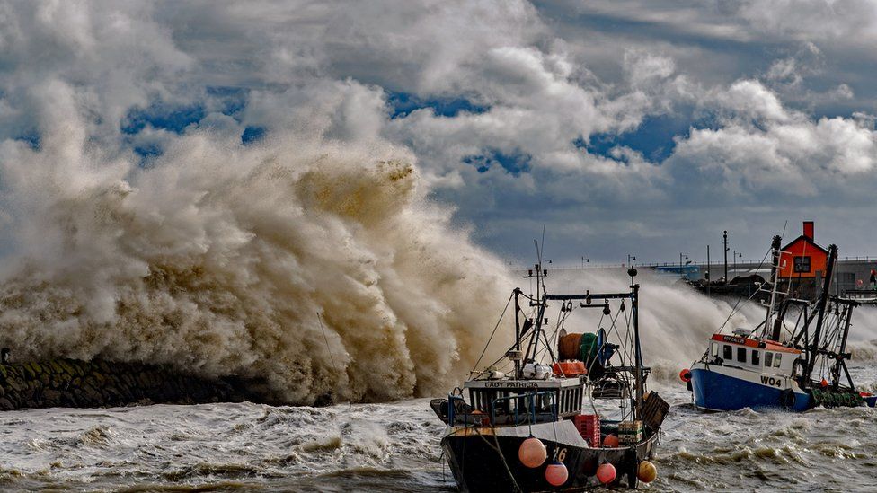 Fishing trawlers in the sea as waves crash against the harbour wall in Folkestone, Kent.